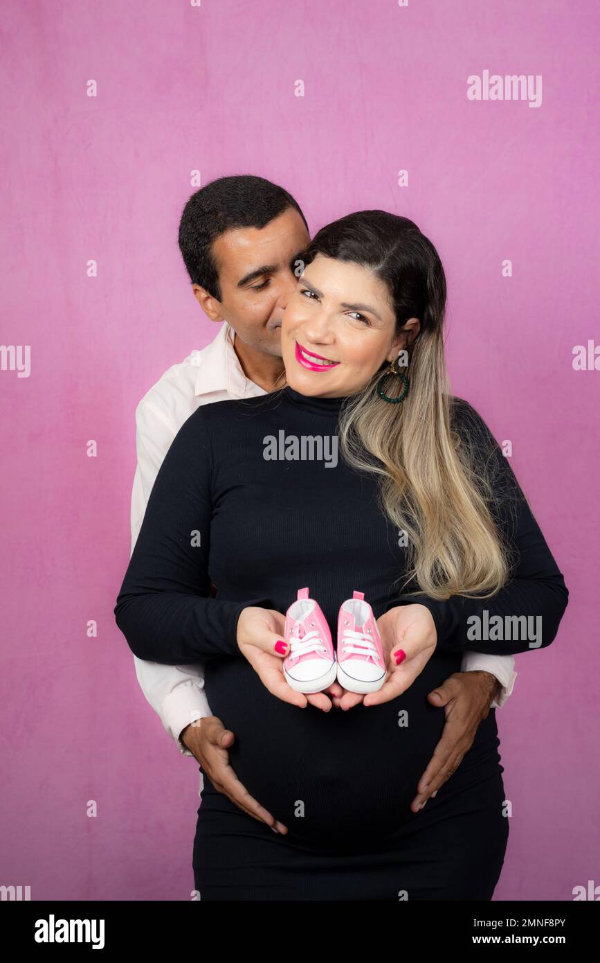 Romantic couple of handsome husband and beautiful pregnant wife posing in studio. She holds baby slippers. Stock Photo