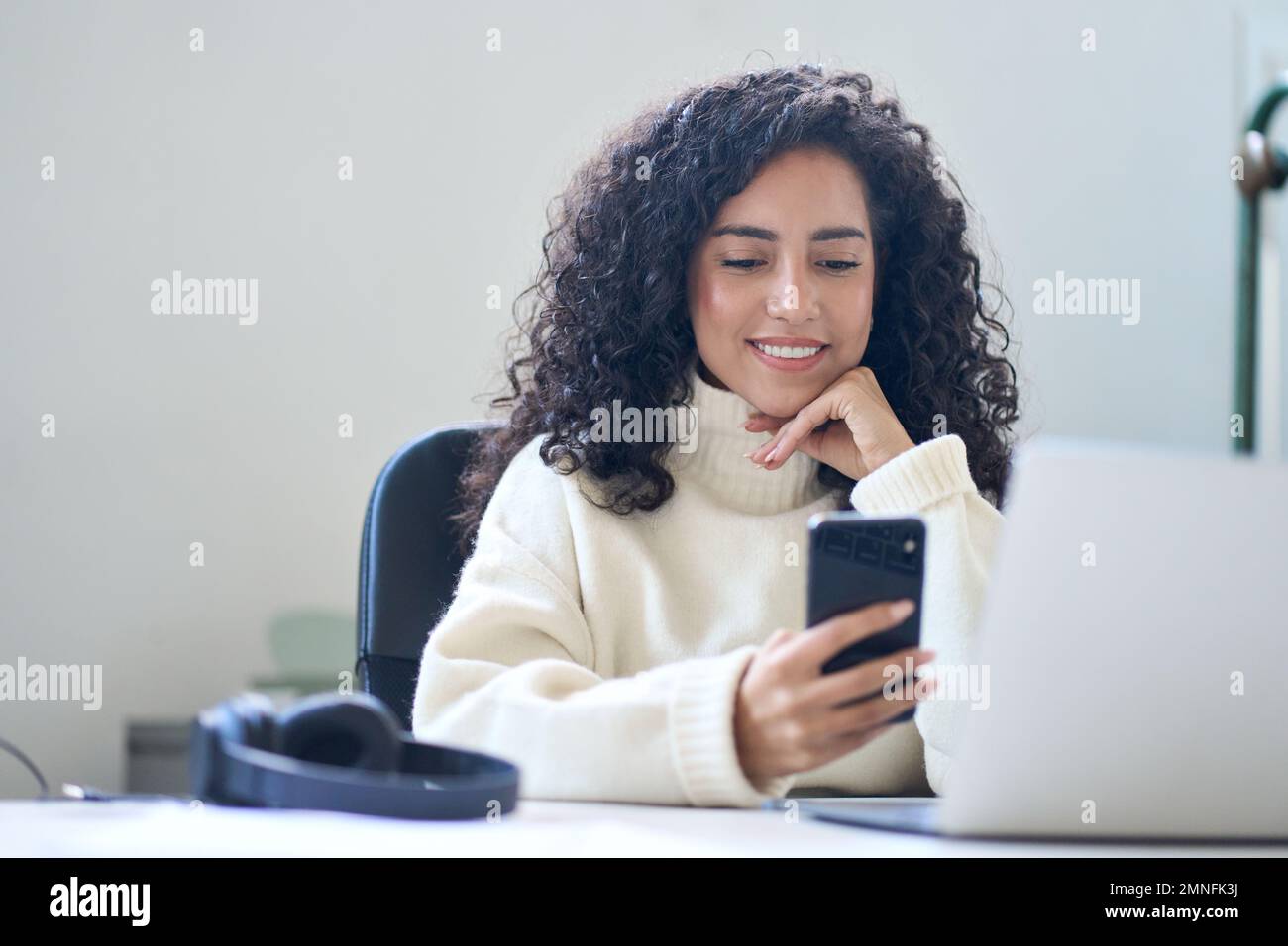 Young smiling latin business woman using phone in office sitting at desk. Stock Photo