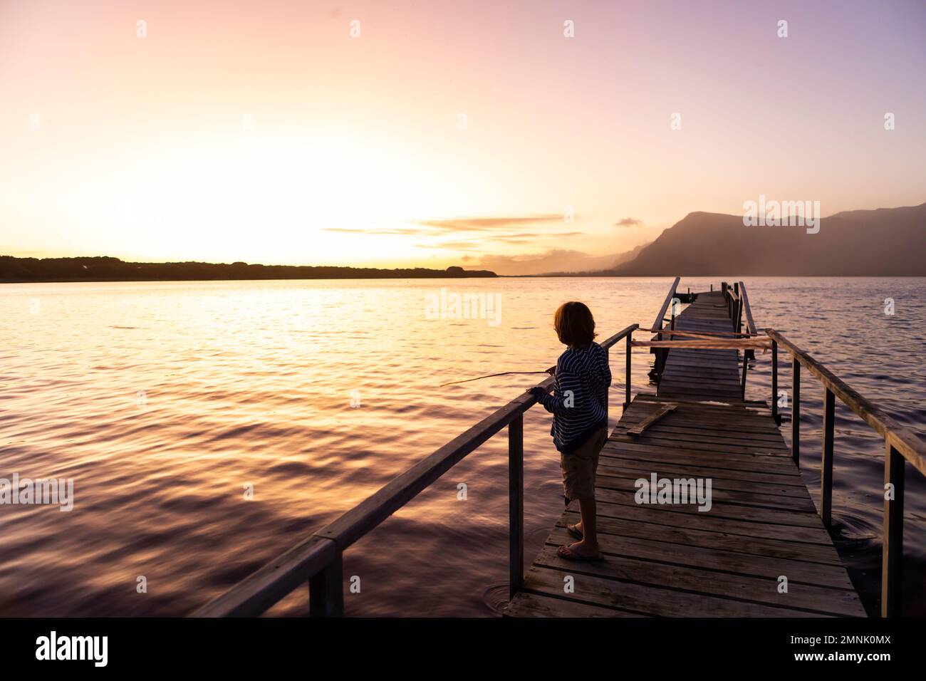 South Africa, Stanford, Boy (10-11) standing on pier at lagoon at sunset Stock Photo