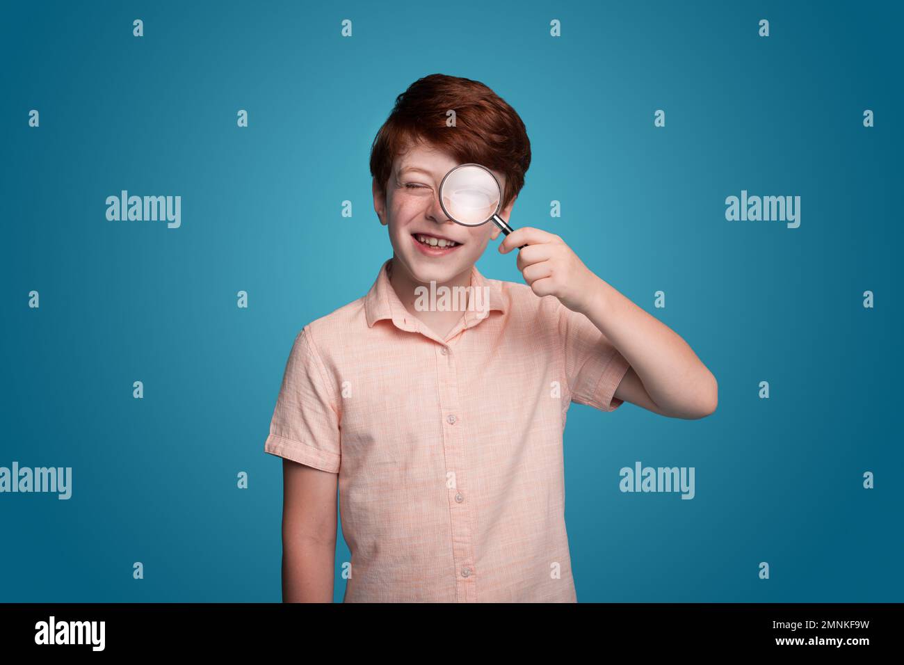 Positive schoolboy in casual wear looking at camera through magnifier while standing against blue background. Education concept. Search concept Stock Photo