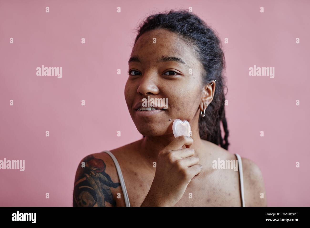 Candid portrait of young black woman with acne scars using face massager looking at camera against pink Stock Photo