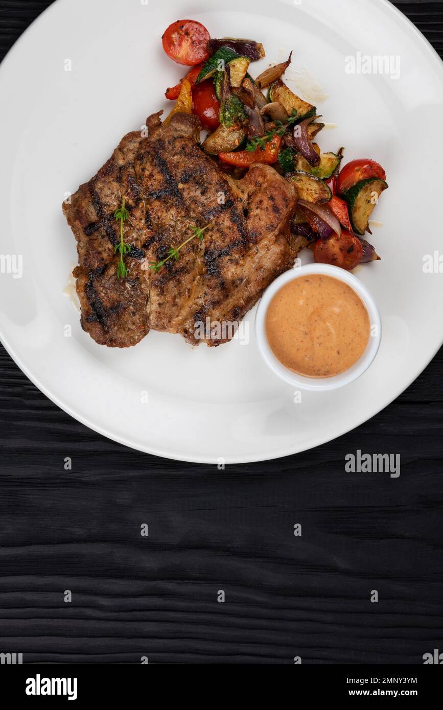 Fried pork chop with grilled vegetables and sauce on white plate on black wooden background Stock Photo