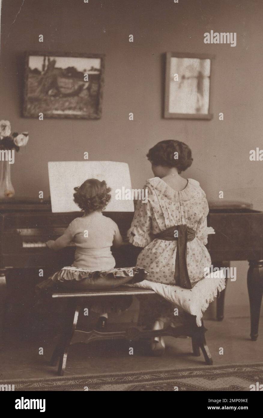 vintage moment / vintage funny moment / vintage photograph / power of the moment / magic moments / vintage piano playing photo. The name of this photo : playing togedher. Little boy or girl is playing on the piano with her/his mother or tutor. Beautiful paintings are hanging on the wall big bunch of flowers on the piano. the photo is from the 1920s. Stock Photo