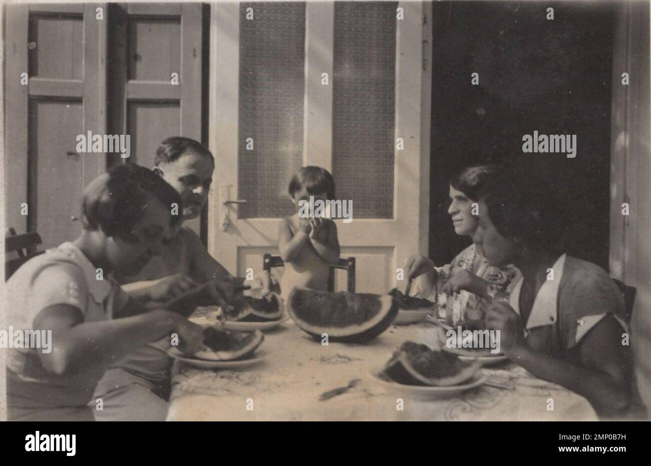 vintage moment / vintage funny moment / vintage photograph / power of the moment / magic moments / vintage eating / eating / vintage photograph about a full family who's are eating nice big watermelon in summer time at the 1930s. The little boy is standing on the chair to reach the nice jucy honey sweet watermelon slice what was the biggest and thats his portion :) Stock Photo