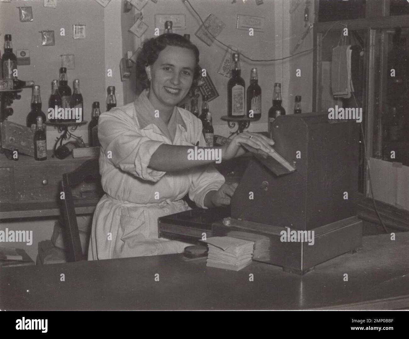 vintage moment / vintage funny moment / vintage photograph / power of the moment / magic moments / happy pretty young lady as cashier behind a vintage cash register  at the 1950s or 1960s. There are lots of bottle of drinks at the backround possibly these bottles are contains alcohol drinks. Stock Photo
