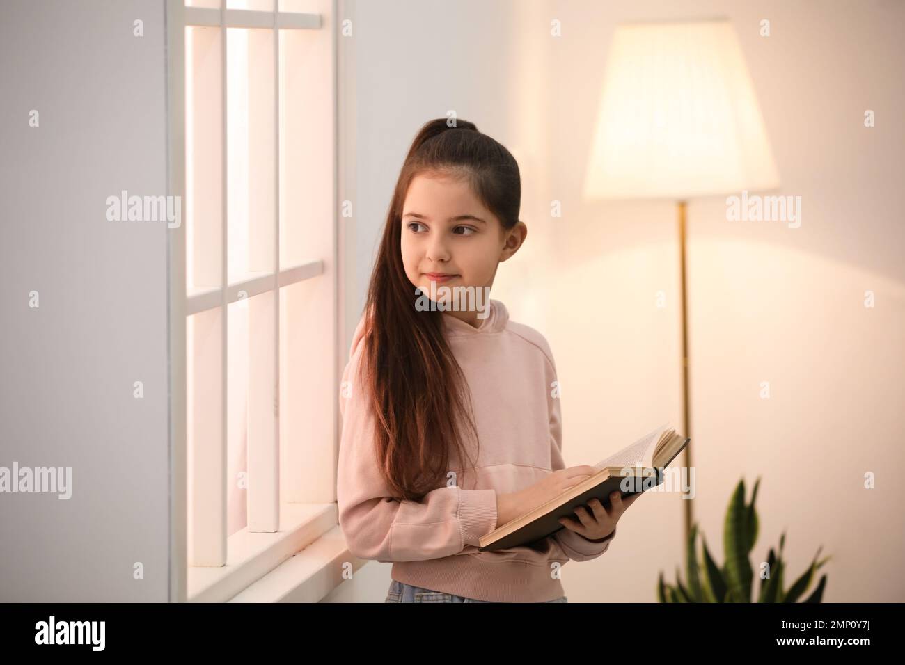 Cute little girl reading book near window at home Stock Photo - Alamy