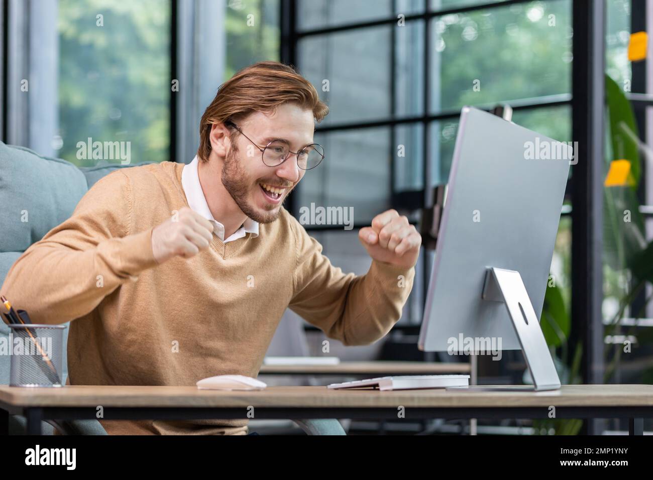 Young man in office celebrating victory and successful achievement results, businessman holding hands in triumph gesture and reading good news from computer , blond programmer in casual clothes. Stock Photo