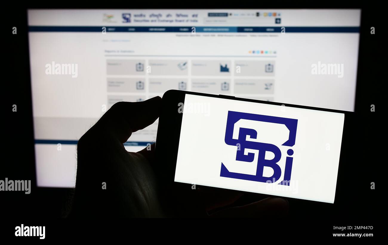 Person holding cellphone with logo of Securities and Exchange Board of India (SEBI) on screen in front of webpage. Focus on phone display. Stock Photo
