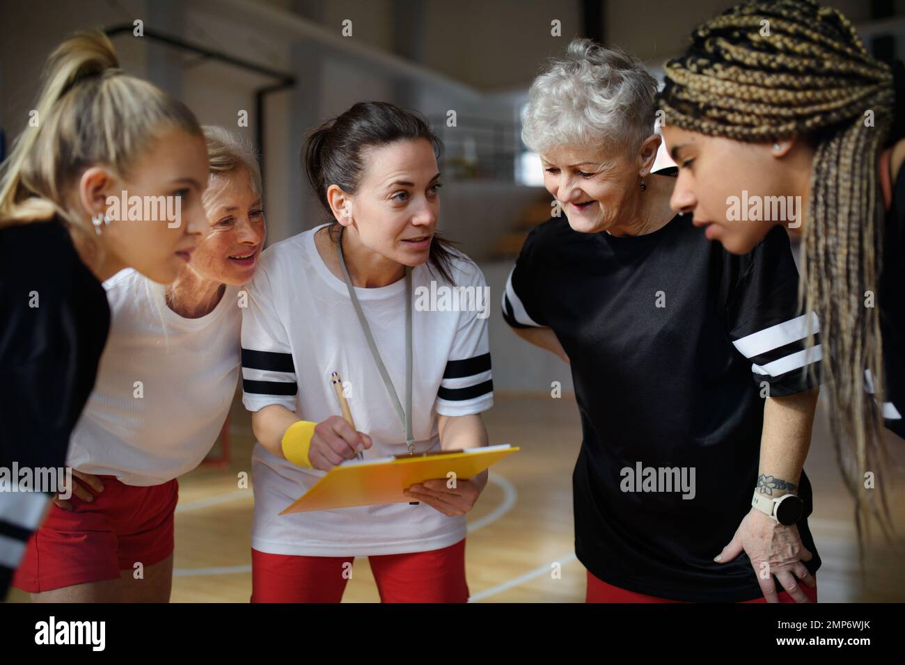 Female sport coach with clipboard discussing tactics with young and old women team training for match in gym. Stock Photo