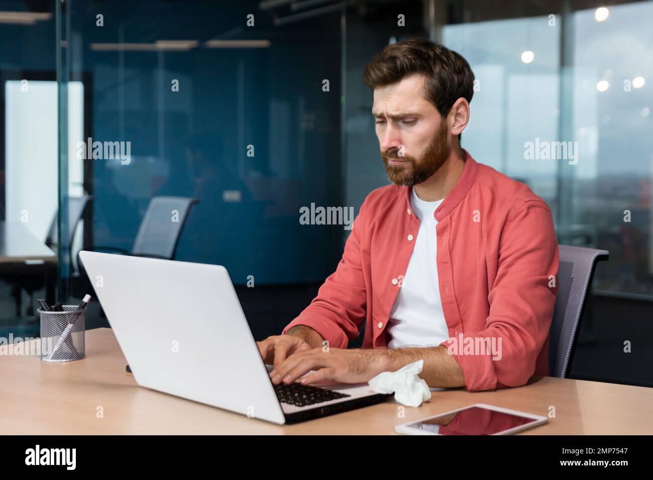 Tired young male designer in a red shirt works in the office at a laptop. He feels bad, got sick, uses a napkin. Stock Photo