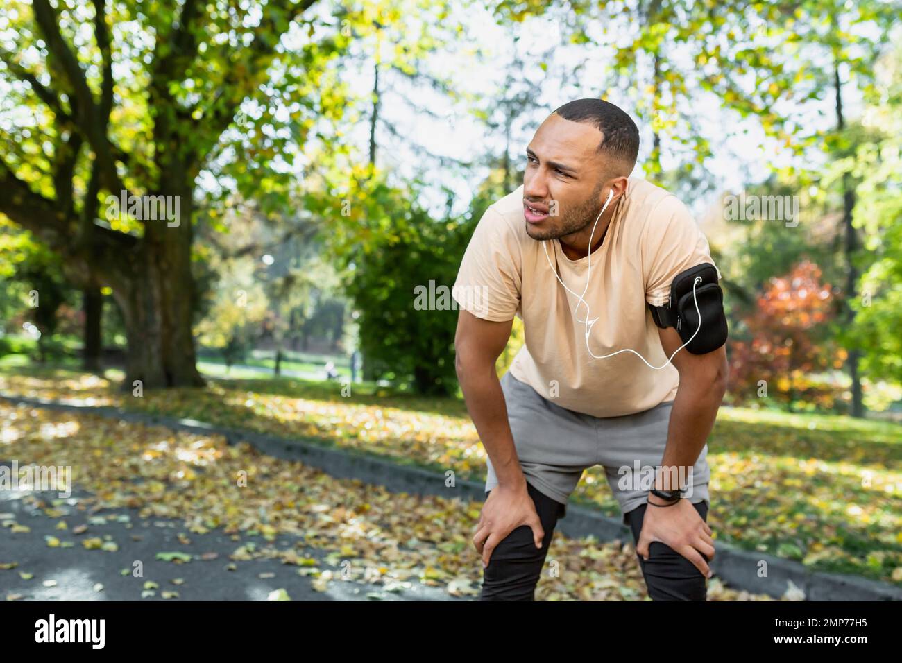 A young African American male athlete listens to music in white headphones and goes for a morning jog in the park. He is tired, stopped, breathes hard, stands and rests. Stock Photo