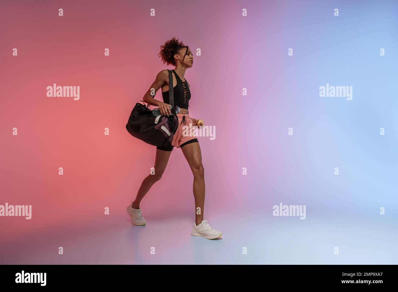 Female athlete holding water bottle, sport bag and apple against studio background  Stock Photo