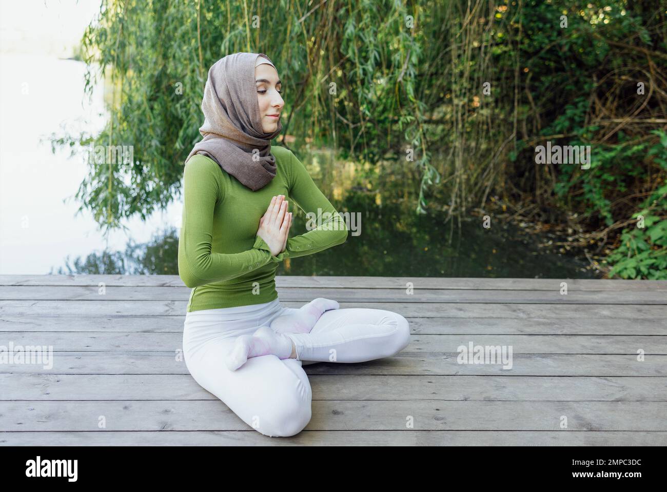 Muslim young girl in a hijab is doing yoga asana in the park. Teenage girl sits in a lotus position on pier by the lake or river. Relax concept Stock Photo