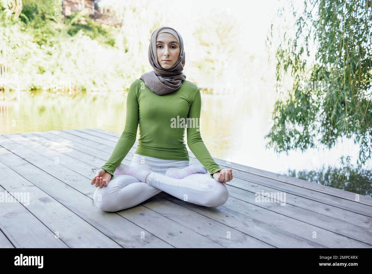 Muslim young girl in a hijab is doing yoga asana in the park. Teenage girl sits in a lotus position on pier by the lake or river. Relax concept Stock Photo