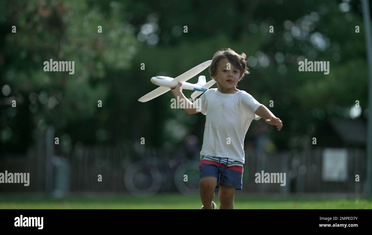 Young boy running with plane toy at park. Carefree Child throwing airplane glider outdoors having fun. Playful kid throws foam plane in sunny beautifu Stock Photo