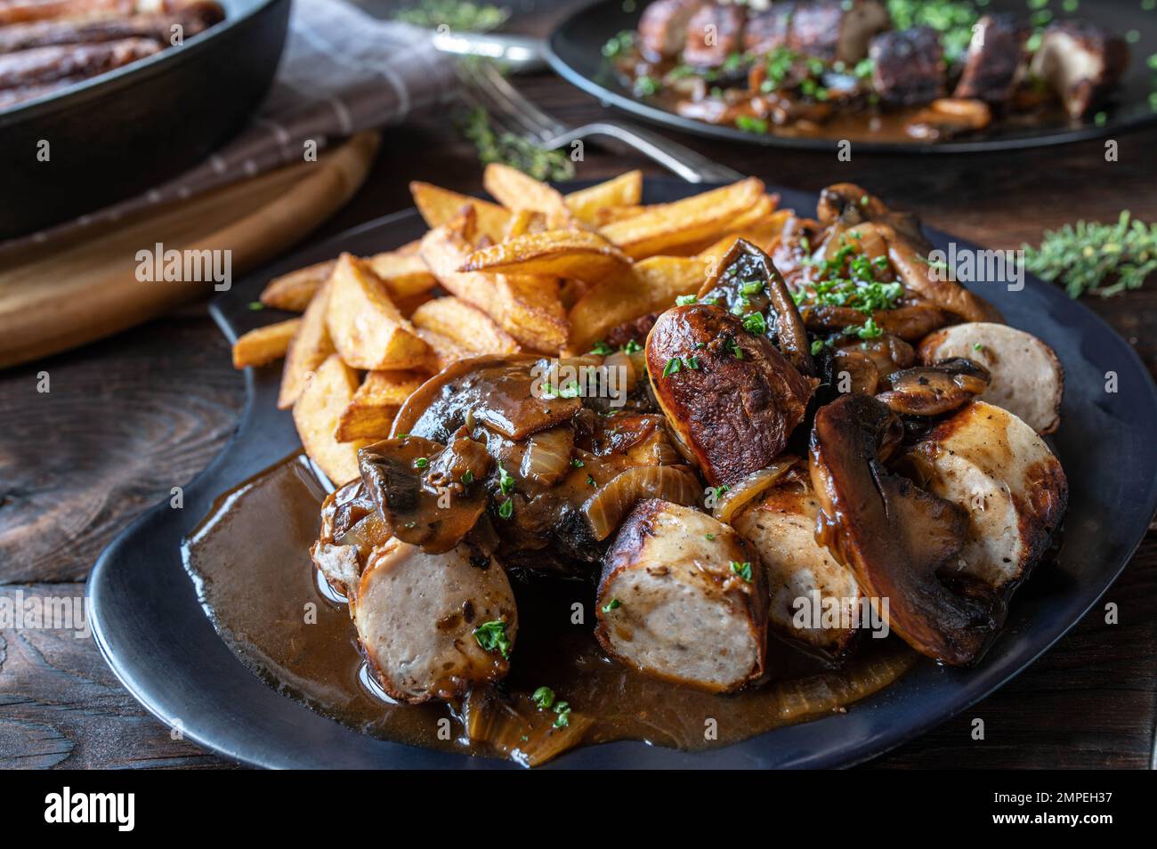 Bratwurst with brown onion, mushroom sauce and homemade french fries on wooden table. Stock Photo