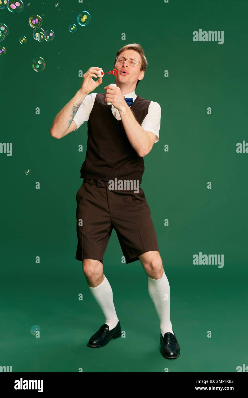 Portrait of man in stylish classical clothes, shorts and vest posing, blowing bubbles over green studio background. Concept of emotions, facial Stock Photo