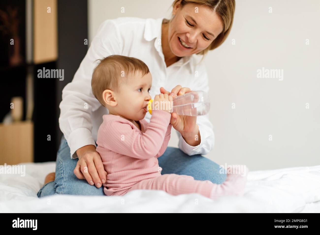Baby hydration. Caring mother giving water bottle to her infant child, sitting on bed at home, copy space Stock Photo