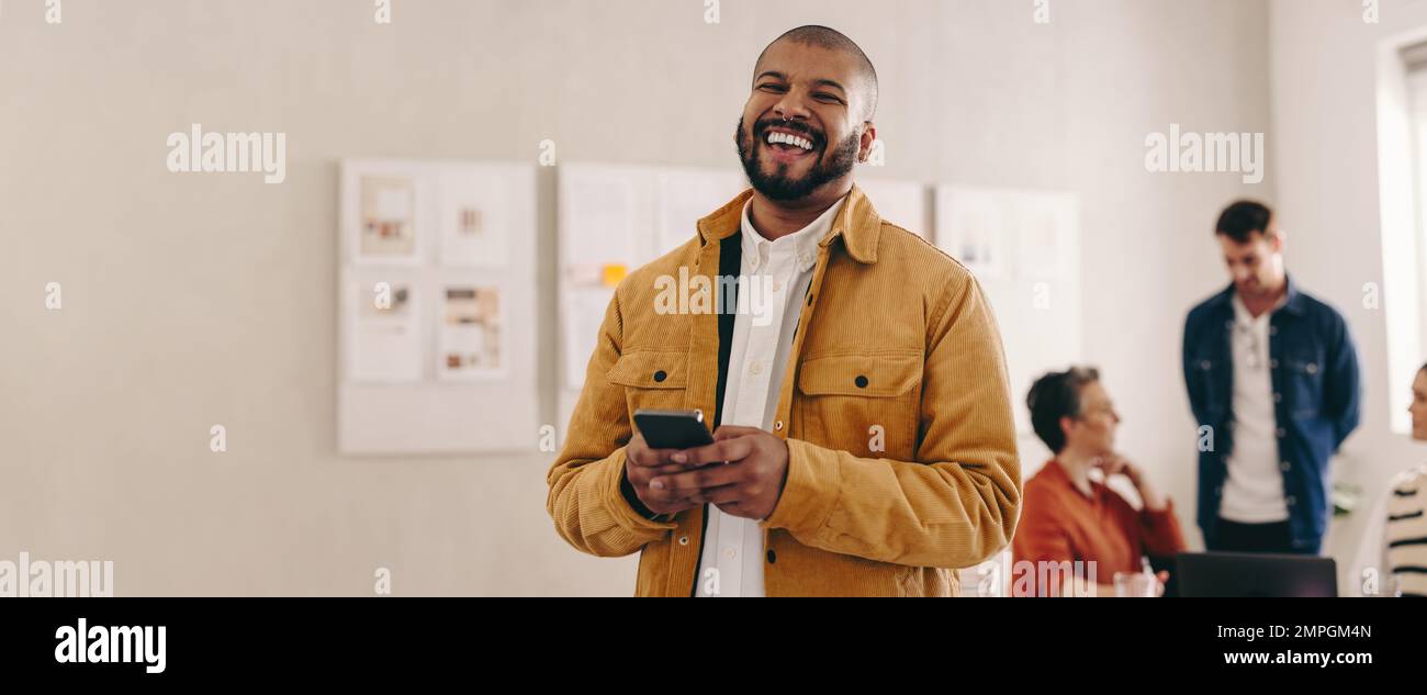 Cheerful businessman with a septum ring smiling at the camera while holding a smartphone. Happy young businessman working in a creative and modern wor Stock Photo