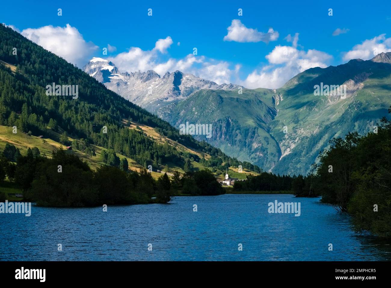 View over the lake Geschinersee to the church of Ulrichen, the snow covered summit of the mountain Galenstock in the distance. Stock Photo