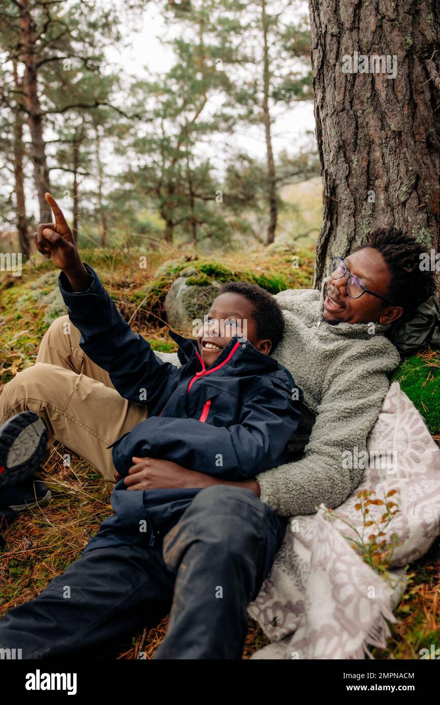 Boy pointing while lying down with father near tree in forest Stock Photo