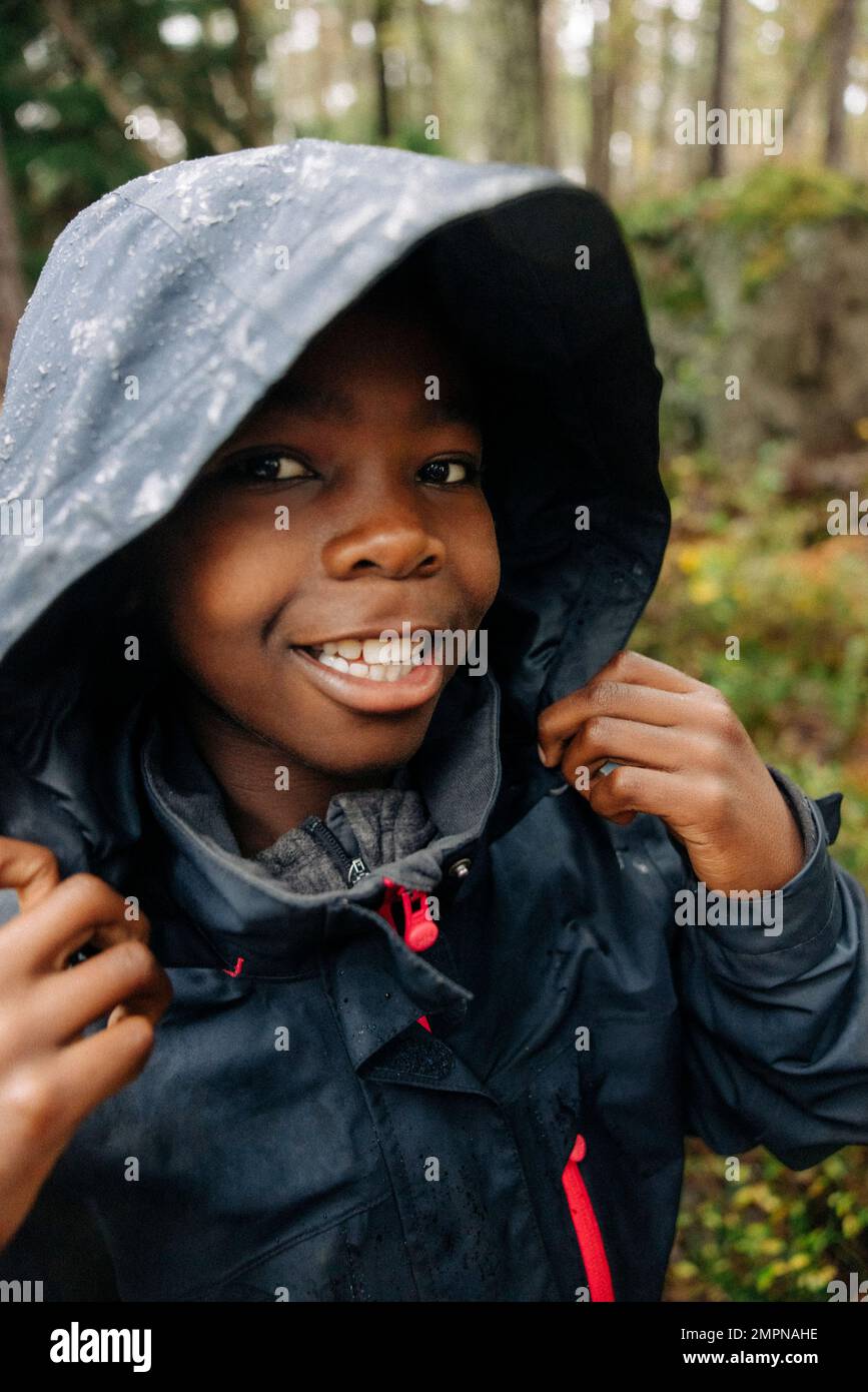 Portrait of boy with toothy smile holding hood of raincoat in forest Stock Photo