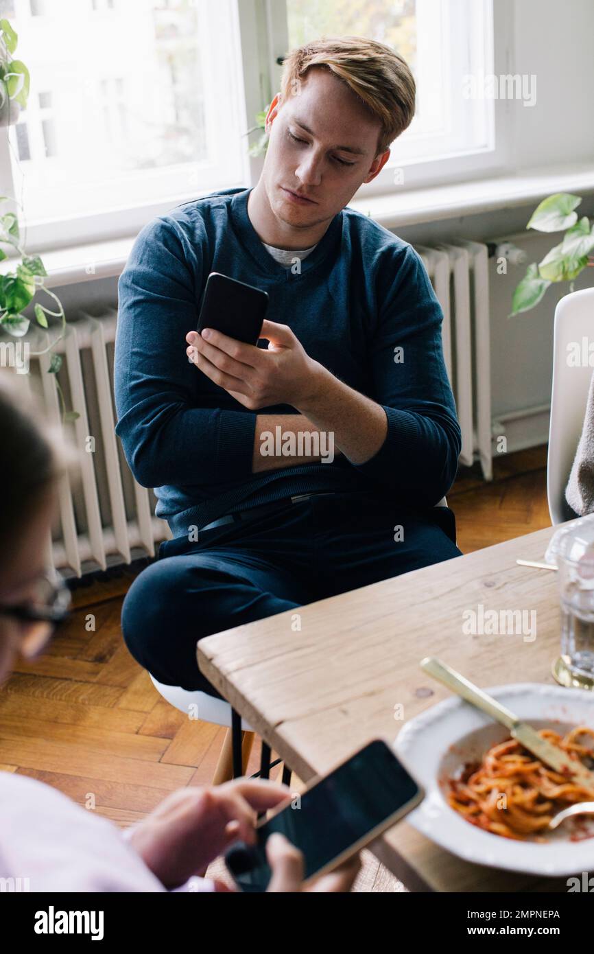 Young man using smart phone sitting at dining table in home Stock Photo