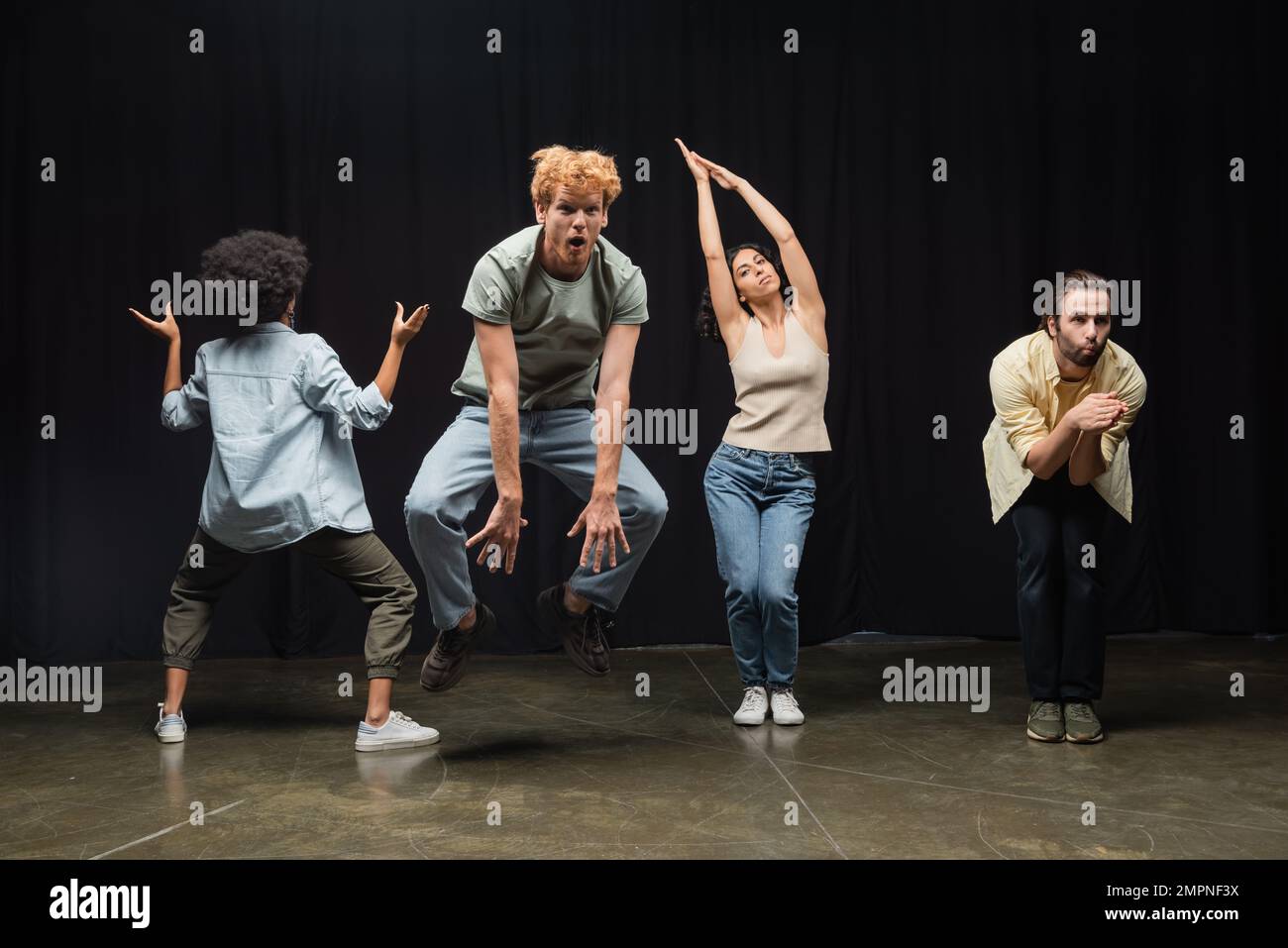excited redhead man jumping near multiethnic actors posing during rehearsal in theater Stock Photo