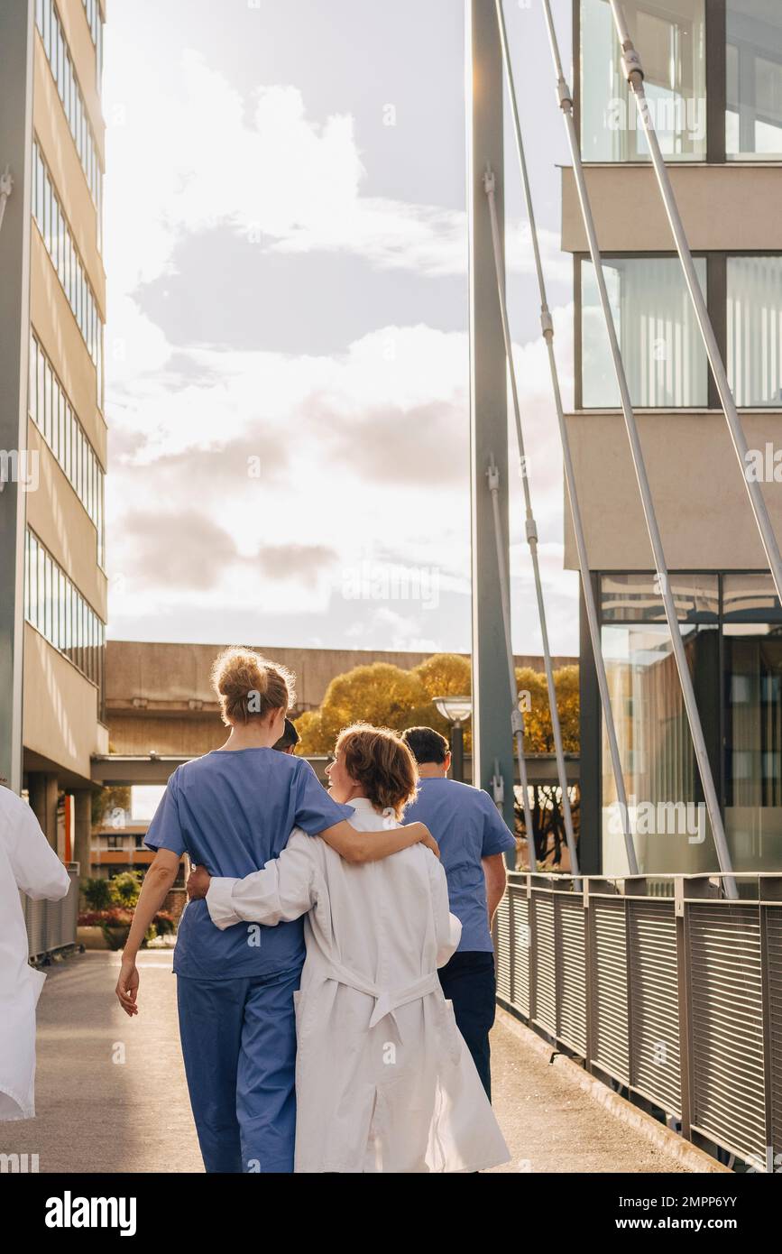Rear view of healthcare workers with arms around each other walking towards hospital during sunset Stock Photo