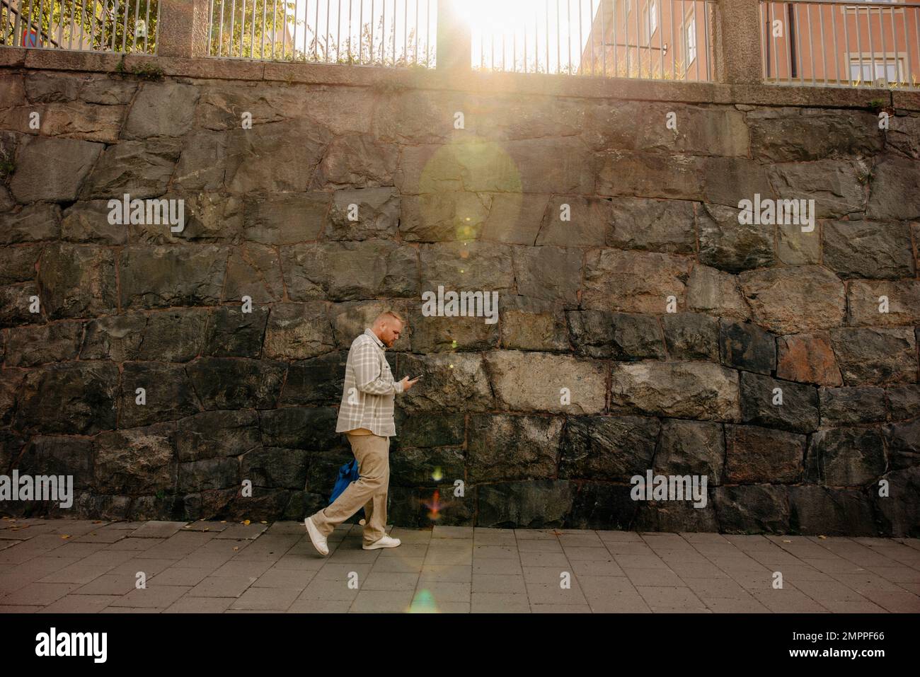 Man using smart phone while walking on footpath in front of wall Stock Photo