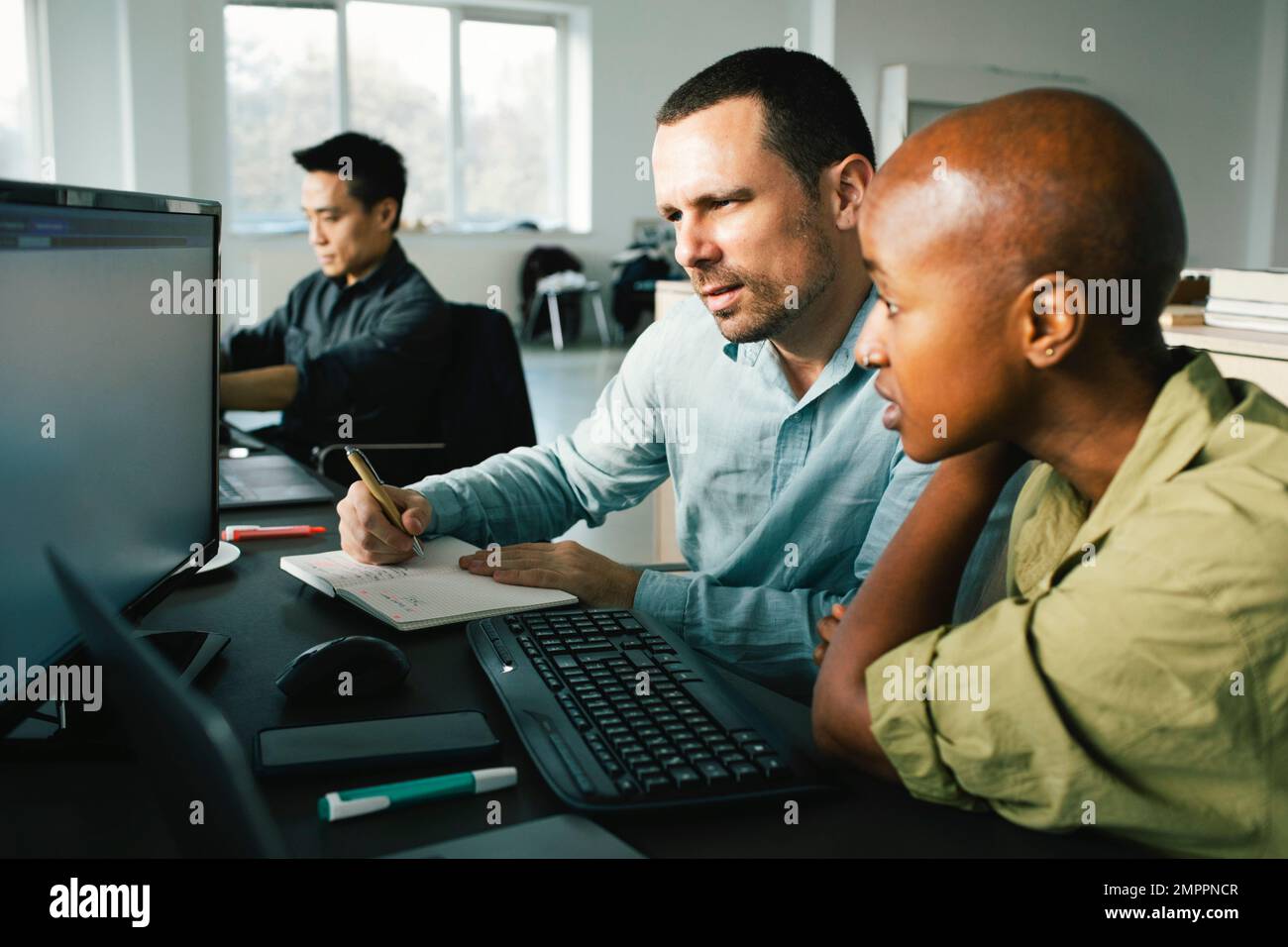 Businesswoman discussing with male colleague taking down notes while looking at computer in office Stock Photo