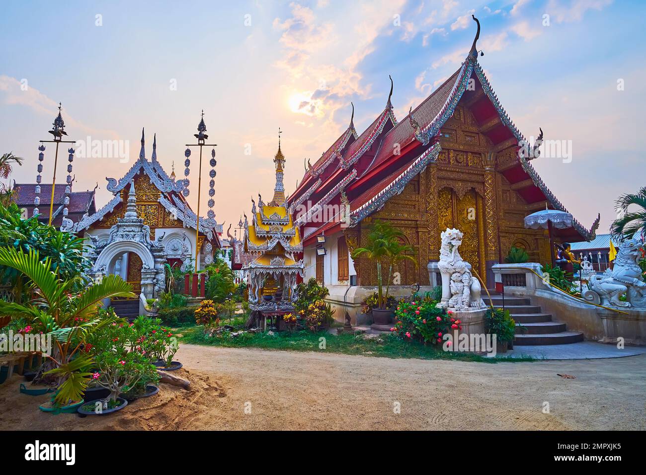 The cloudy sunset sky over the ornate shrines of medieval Wat Mahawan, surrounded with flowering garden, Chiang Mai, Thailand Stock Photo