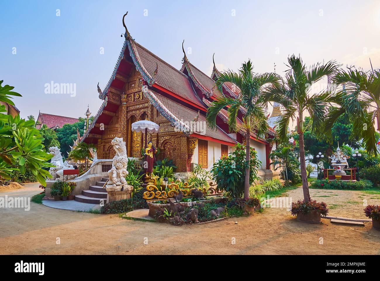 The tropic green garden in Wat Mahawan with medieval sculptured Viharn behind the palms, Chiang Mai, Thailand Stock Photo