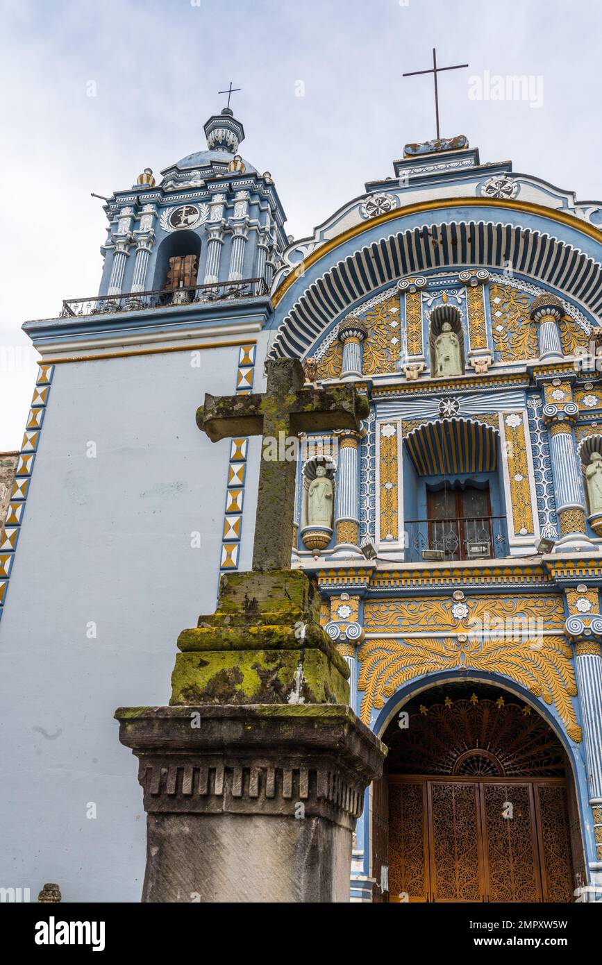 The colorful painted facade and bell tower of the Church of Santo Domingo de Guzman in Ocotlan de Morelos, Oaxaca, Mexico, with a stone cross in front Stock Photo