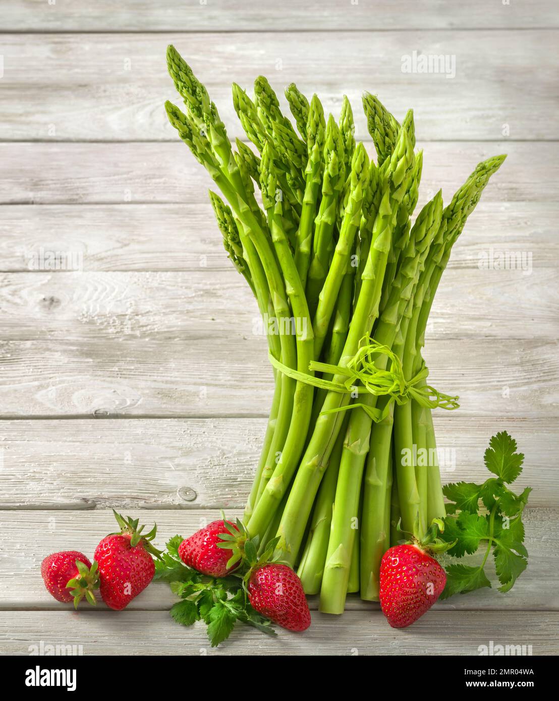 Bunch of green raw asparagus decorated with fresh strawberries on bright pale wood background, vertical format Stock Photo