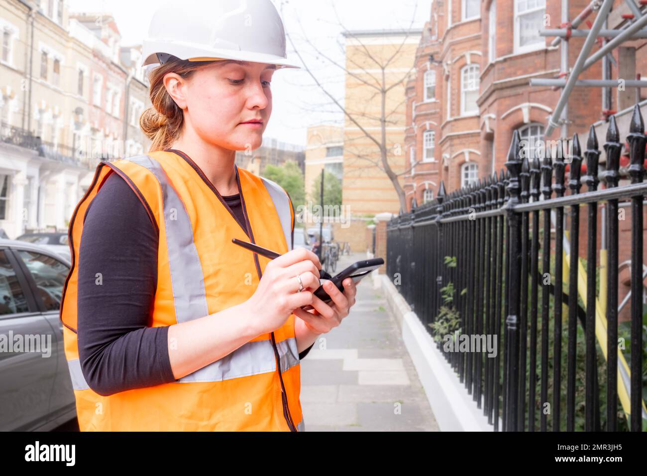 Young adult chartered civil engineer woman taking notes on her smart phone with an electronic pen, construction site, hard hat and orange personal pro Stock Photo