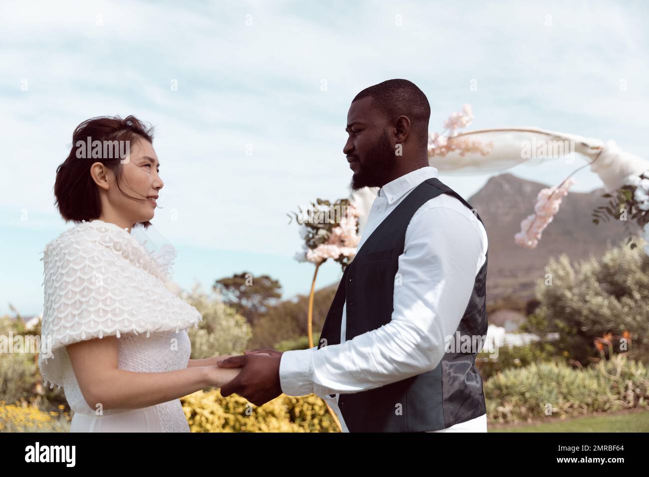 Happy, diverse bride and groom holding hands and looking at each other ...