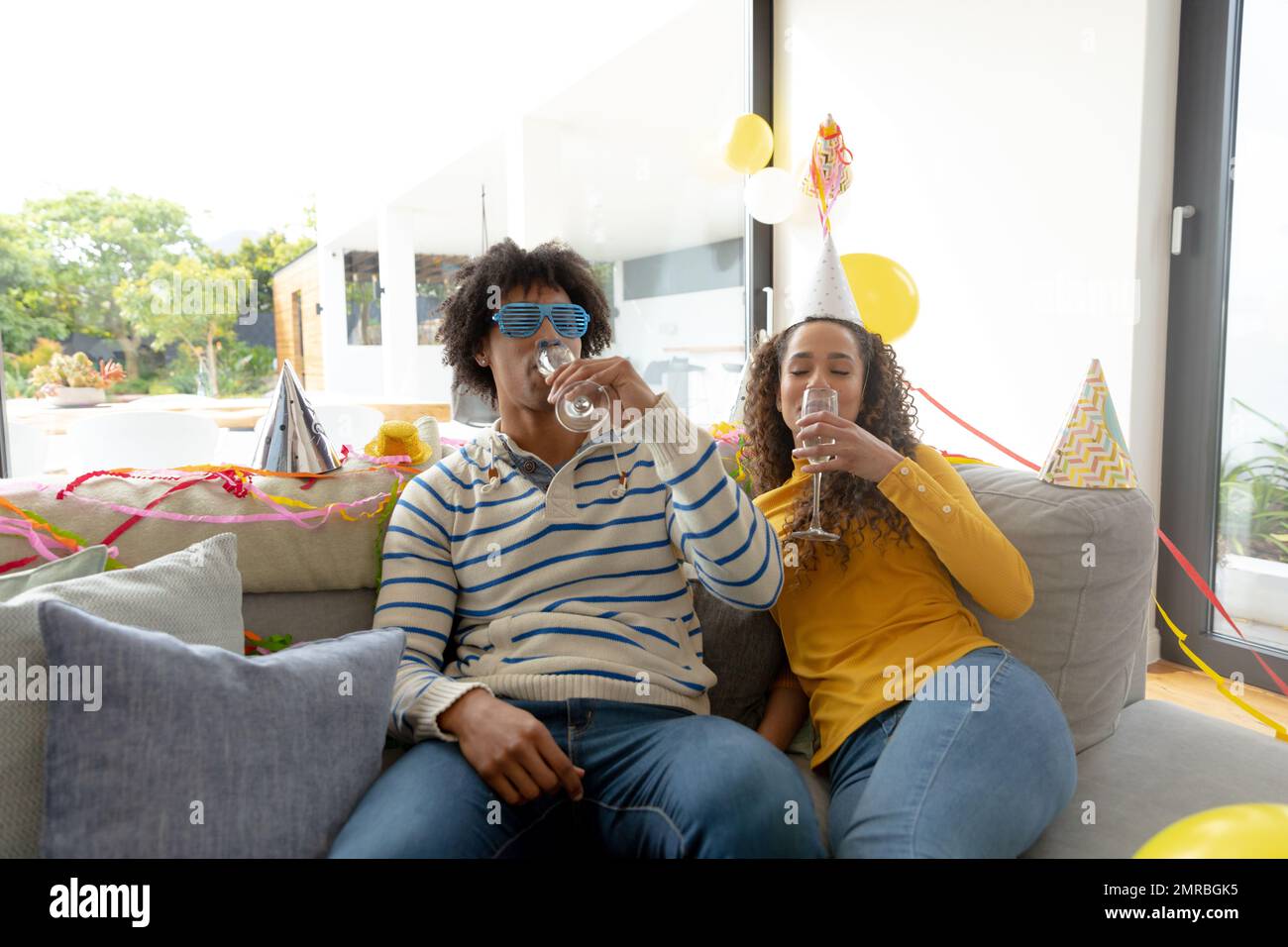 Happy biracial couple having party at home, sitting on couch drinking