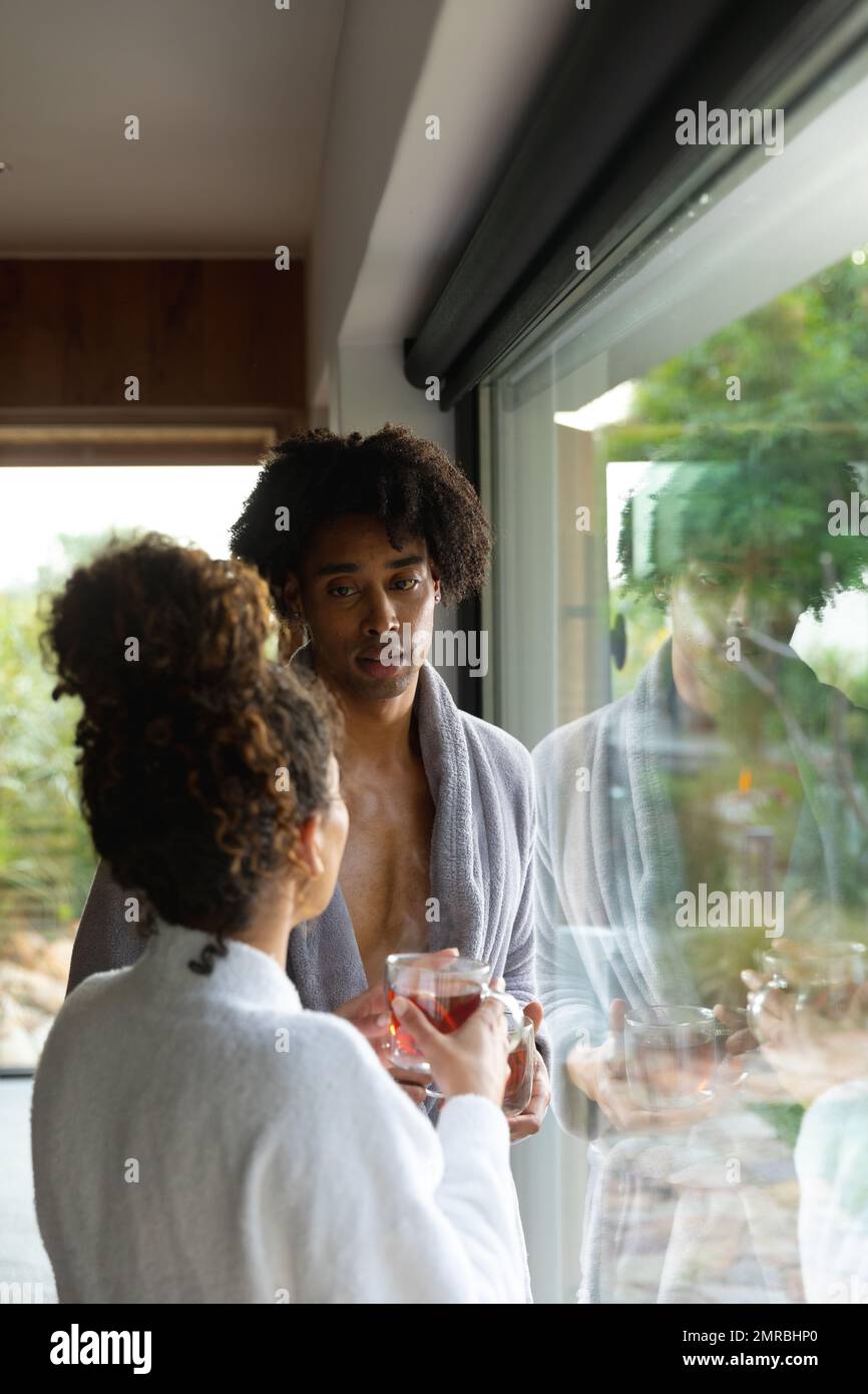 Vertical of biracial couple wearing bathrobes, drinking tea and talking at health spa, copy space Stock Photo