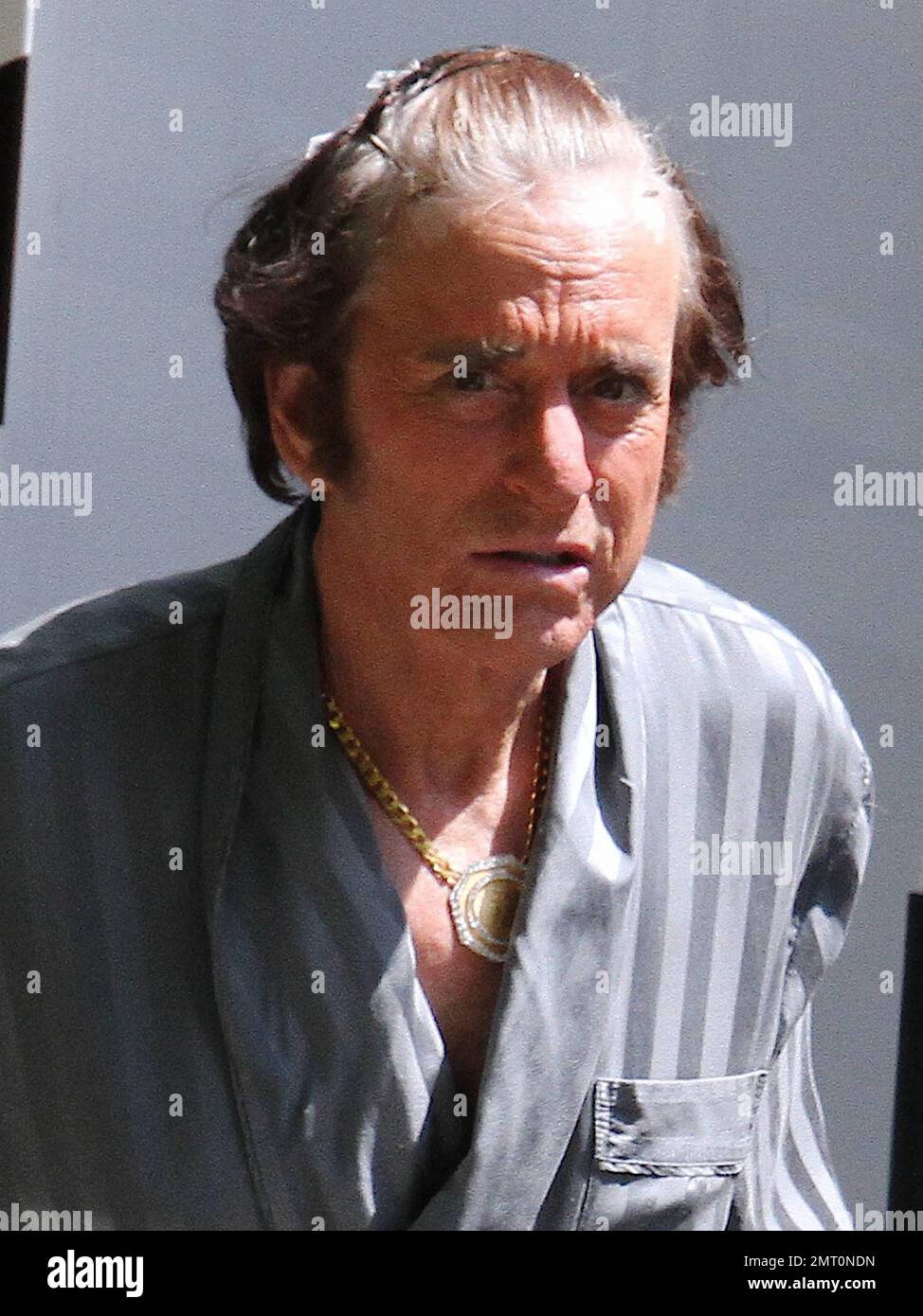 EXCLUSIVE!! Looking very much the character, Michael Douglas wears his hair pinned back from his face while on the set of 'Liberace: Behind the Candelabra.' Douglas, who wore a large gold medallion necklace beneath his grey robe, looked heavily tanned like his character. He also scratched lightly at the clips holding his hair in place as he walked on set. In the film, Douglas plays Liberace and Matt Damon plays his boyfriend, Scott Thorson. Palm Springs, CA. 20th August 2012. Stock Photo