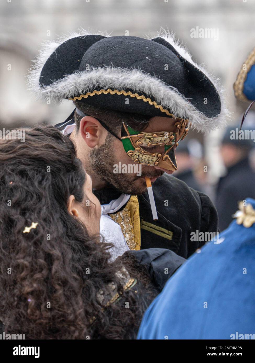 Handsome young man in carnival mask wearing tricorn with an unlit cigarette in his mouth at Venice carnival among his friends Stock Photo