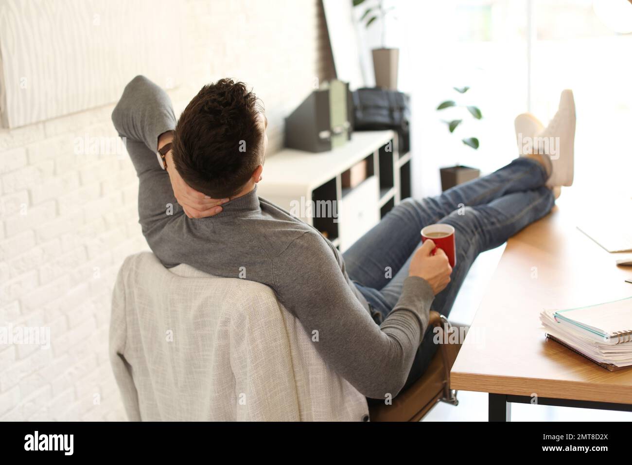 Young man with cup of drink relaxing at table in office during break Stock Photo