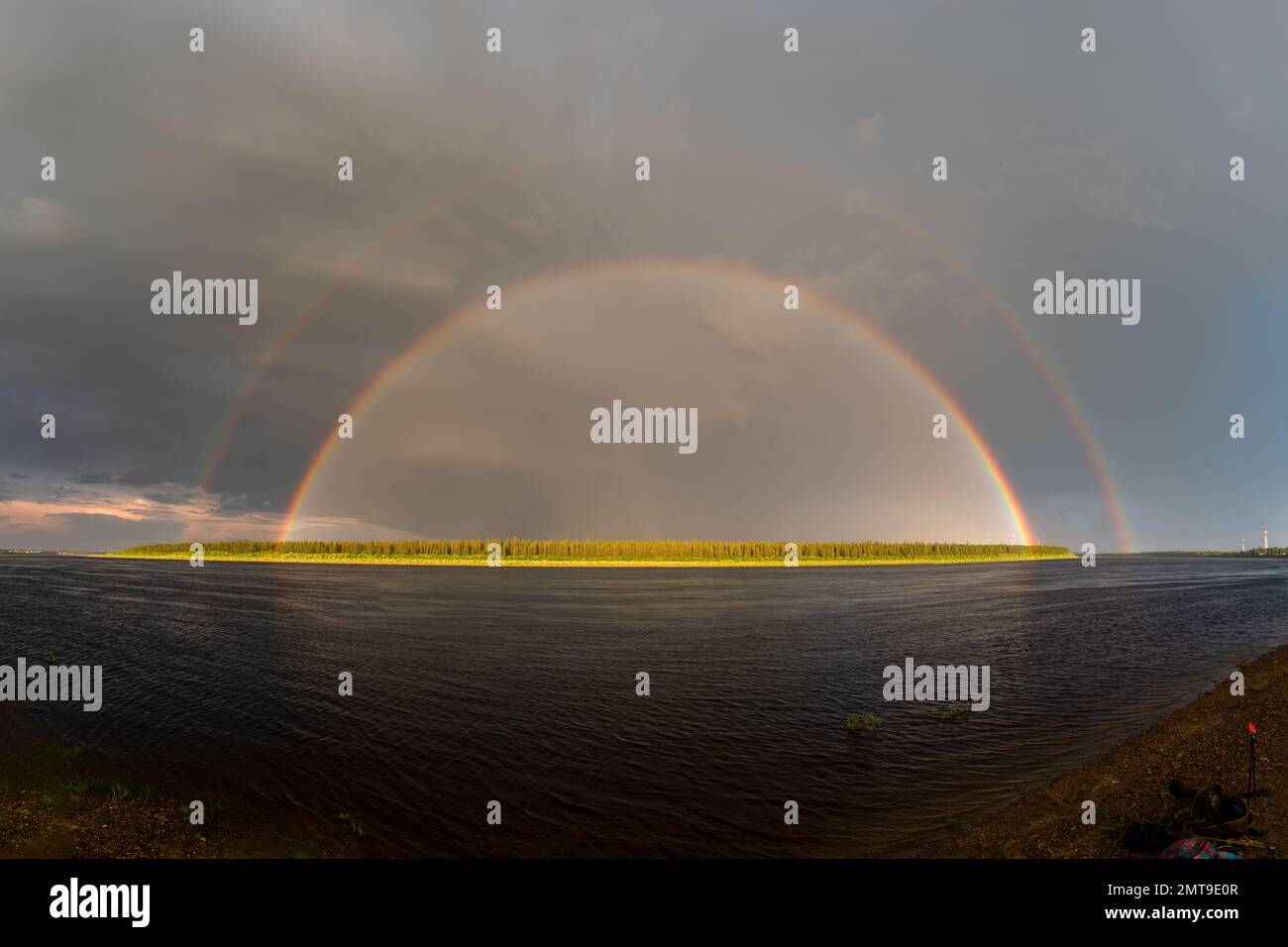 A panorama of a double rainbow against the backdrop of a bright sky emerges from the pylons of power lines and illuminated by the forest on the banks Stock Photo