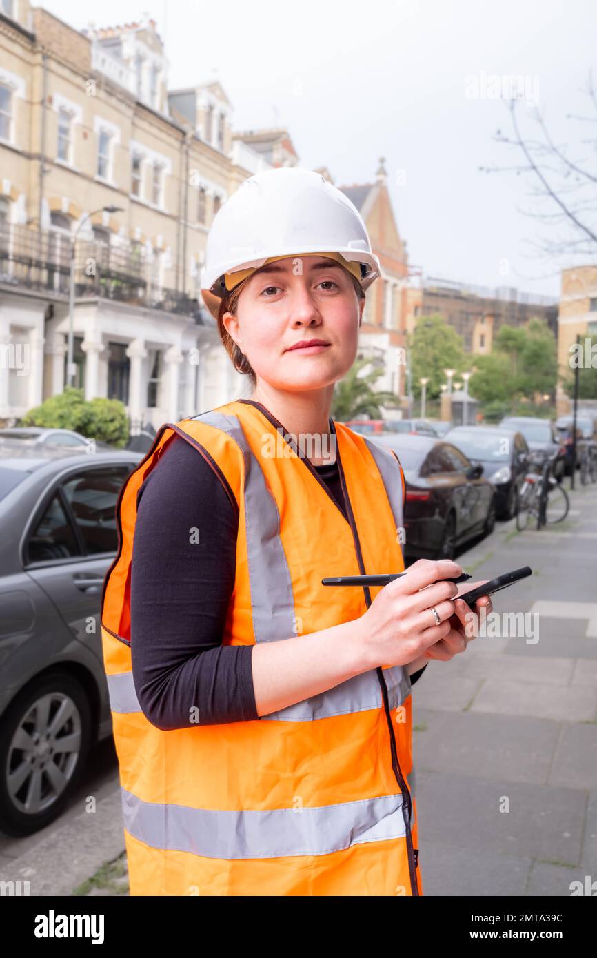 Female chartered civil engineer writing on a smart phone with electronic pen, construction site, hard hat and orange personal protective equipment, co Stock Photo