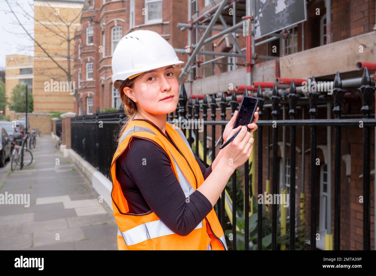 Chartered civil engineer lady showing you something on a smart phone with electronic pen, construction site, hard hat and orange high visibility vest, Stock Photo