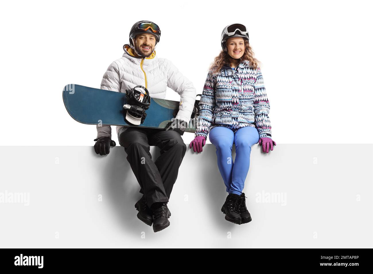 Young man and woman wearing winter equipment sitting on a blank panel and holding a snowboard isolated on white background Stock Photo