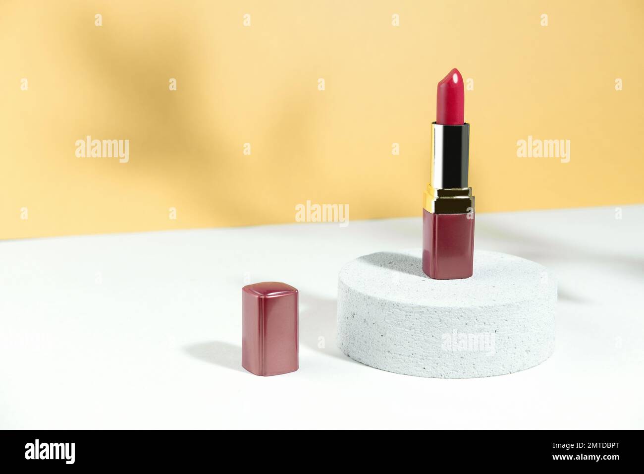 A tube of red lipstick on the podium, on a white orange autumn background, with a tough shadow from the plant. Protects and nourishes the lips. Contai Stock Photo