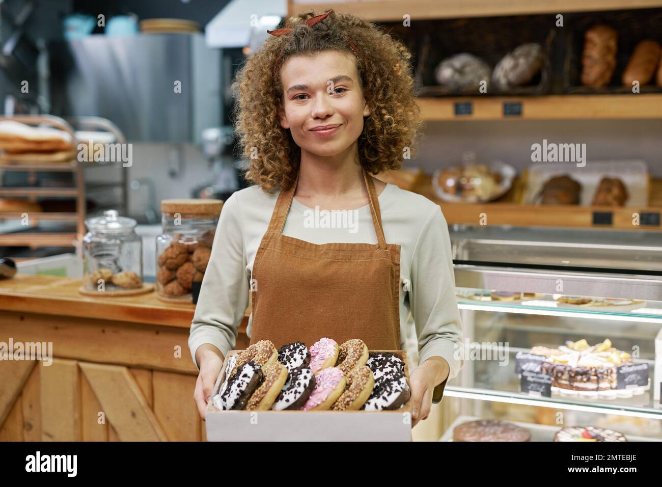 Portrait of cheerful young bakery owner holding tray of fresh glazed donuts Stock Photo