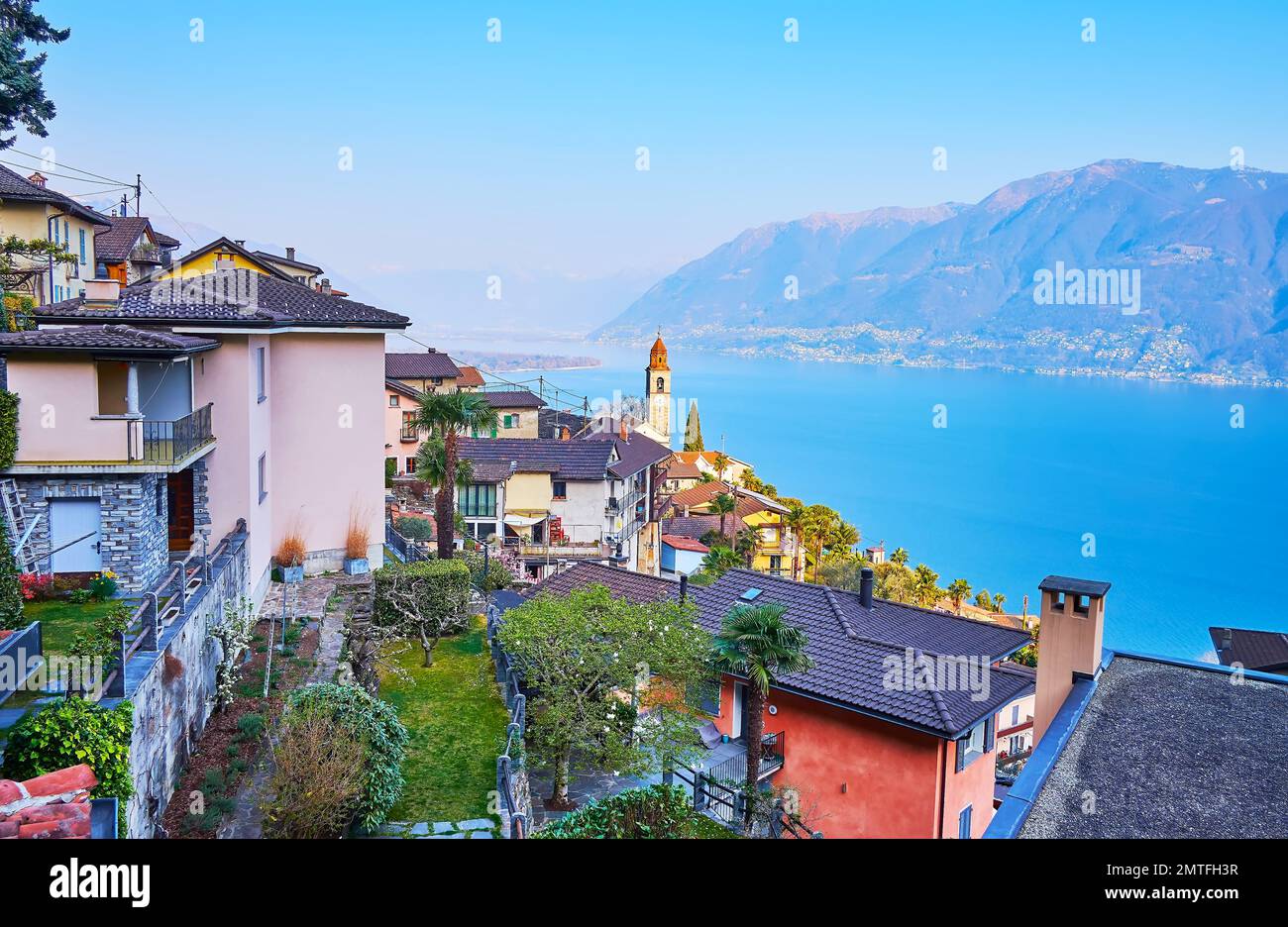 The beautiful mountain village of Ronco sopra Ascona with small houses, green gardens, clocktower of San Martino Church and blue Lake Maggiore in back Stock Photo