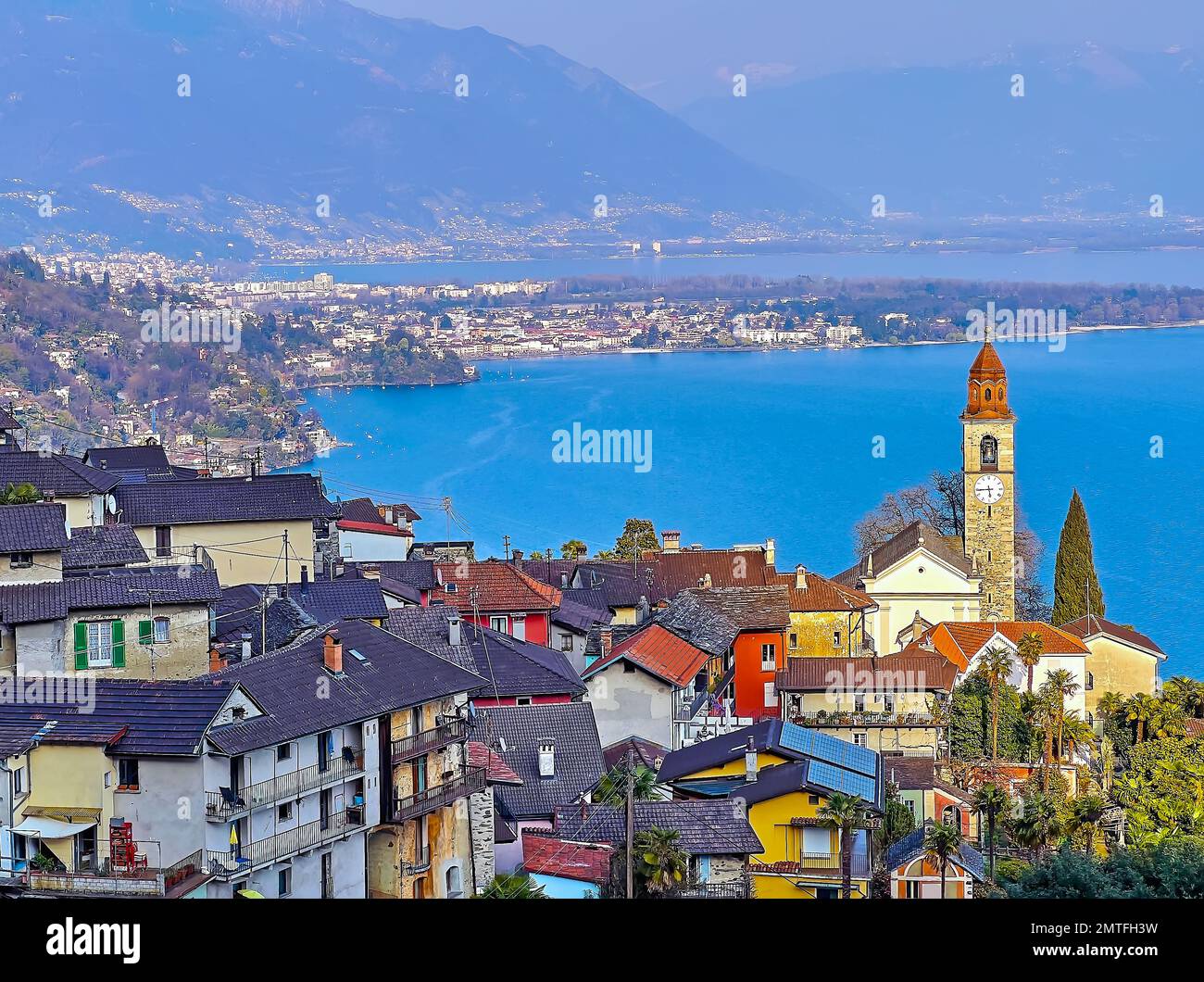 Slender bell tower of San Martino Church and vintage houses of Ronco sopra Ascona against the hazy Lake Maggiore and Alpine slopes, Switzerland Stock Photo